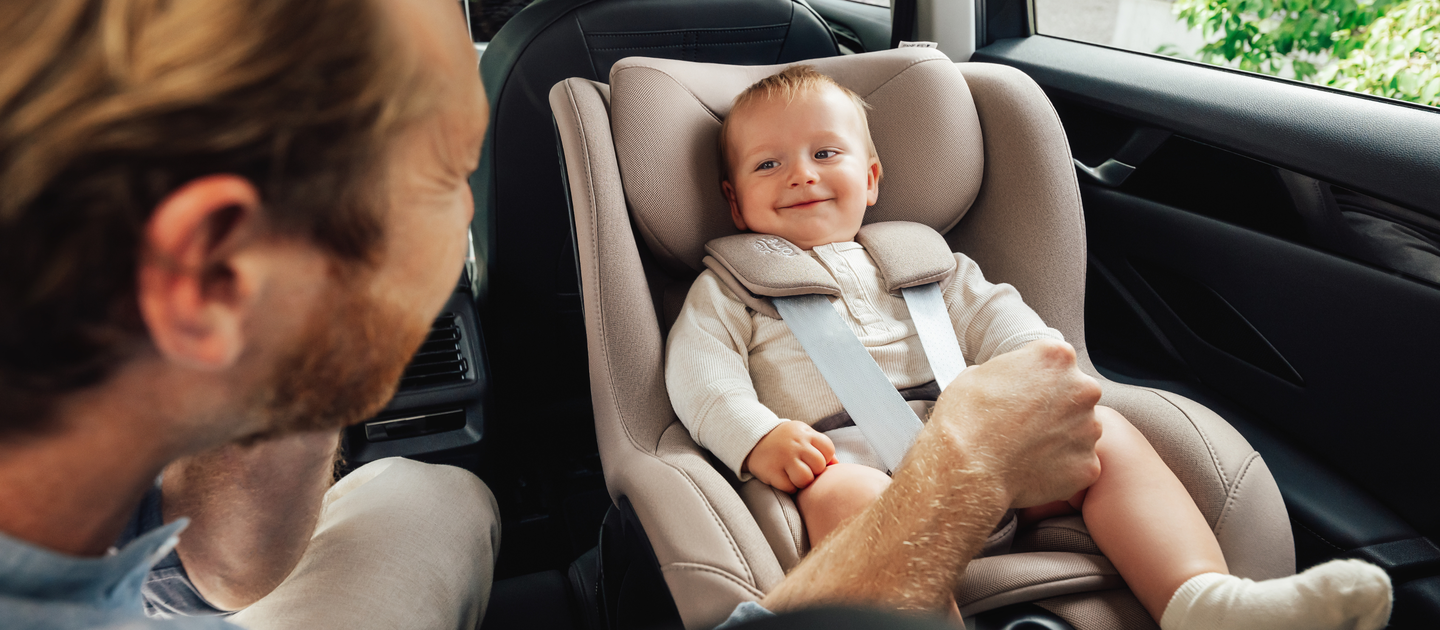 Un b&eacute;b&eacute; souriant est assis en toute s&eacute;curit&eacute; dans un si&egrave;ge auto Britax R&ouml;mer Dualfix 5Z couleur Teak, install&eacute; sur la banquette arri&egrave;re d&rsquo;une voiture. Un adulte, vu de c&ocirc;t&eacute;, tient doucement la jambe du b&eacute;b&eacute; tout en interagissant avec lui.